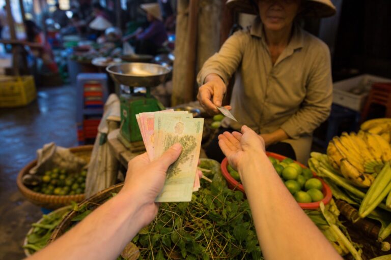 A traveller paying a Vietnamese market vendor with dong banknotes while she hands back change at a produce stall