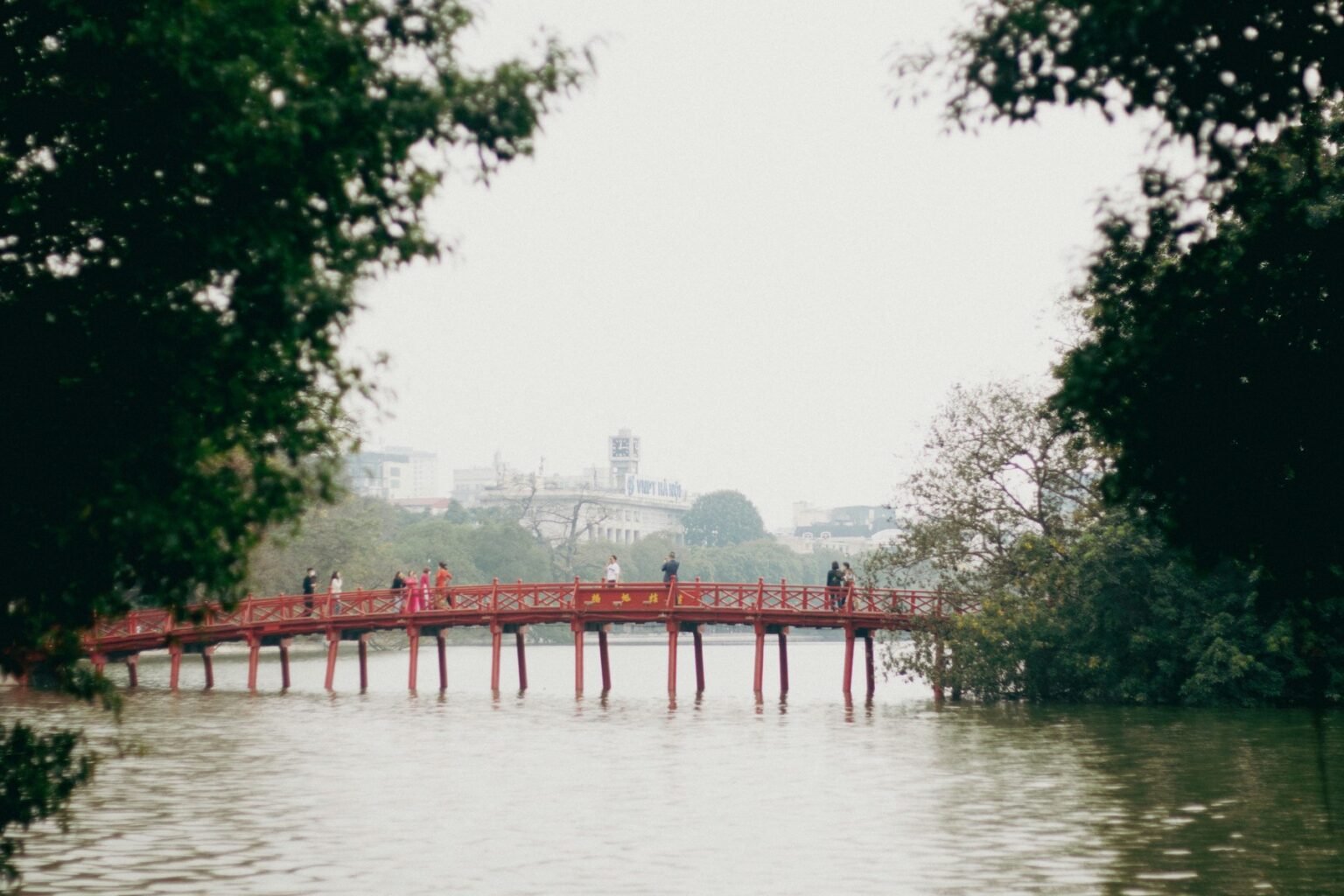 Red The Huc Bridge over Hoan Kiem Lake in Hanoi, framed by green trees